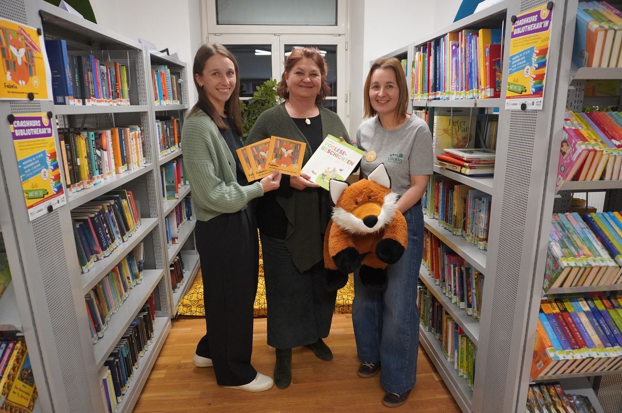 Drei Frauen stehen in einer Bibliothek mit Bücherregalen, halten Bücher und einen pelzigen Fuchs und lächeln in die Kamera.