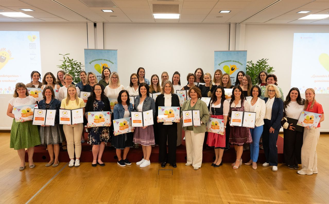 Eine Gruppe von Frauen mit Urkunden steht zusammen in einem Raum mit Bannern und Topfpflanzen. Sie scheinen eine Errungenschaft zu feiern.