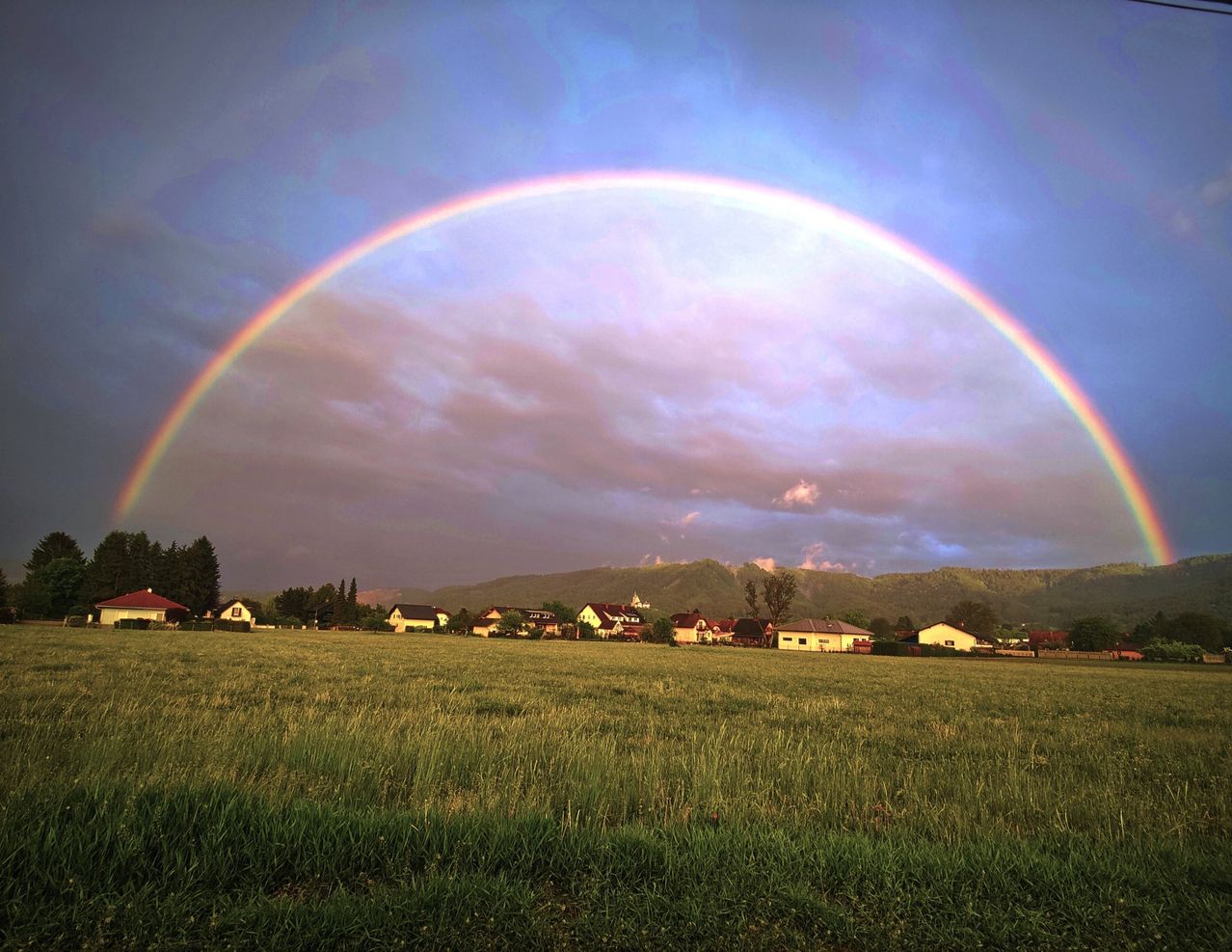 Ein leuchtender Regenbogen erstreckt sich über eine ruhige Landschaft mit Häusern und einem Hintergrund aus Bergen unter einem teilweise bewölkten Himmel.