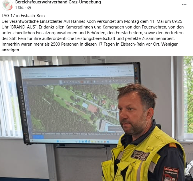 Ein Mann in einer gelben Weste steht vor einem Monitor, der eine Karte anzeigt, wahrscheinlich ein Feuerwehrmann. Er schaut in die Kamera und der Monitor zeigt eine Karte mit einigen grünen Flächen und Gebäuden.