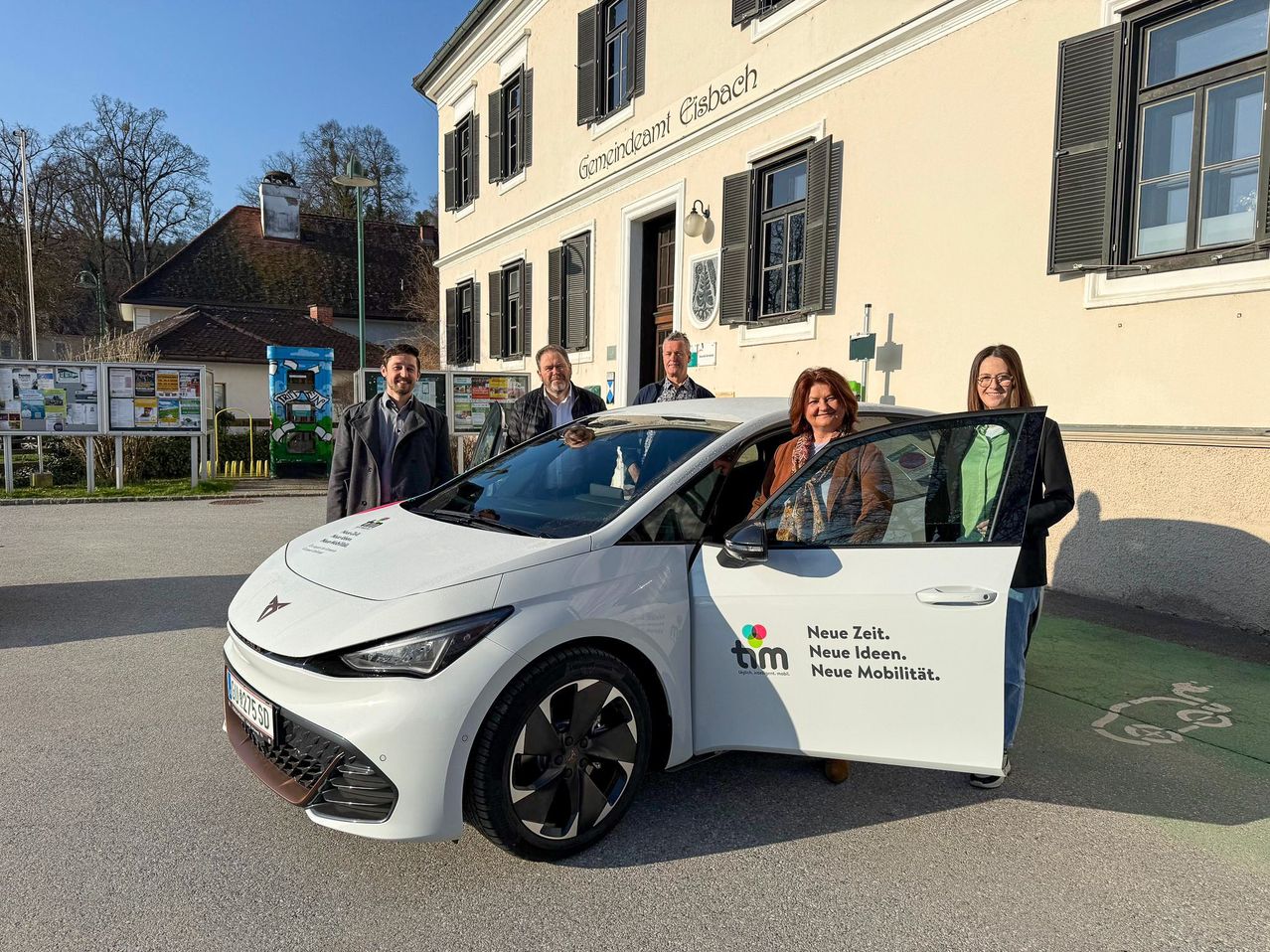 Fünf Personen stehen neben einem weißen Auto mit 'Neue Zeit. Neue Ideen. Neue Mobilität.' davor, vor einem Gebäude.