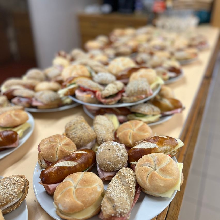 Ein Tisch mit mehreren Platten mit Sandwiches, einschließlich Brötchen, Schinken, Käse und Salat. Die Sandwiches sind in Reihen angeordnet, und einige sind halbiert.