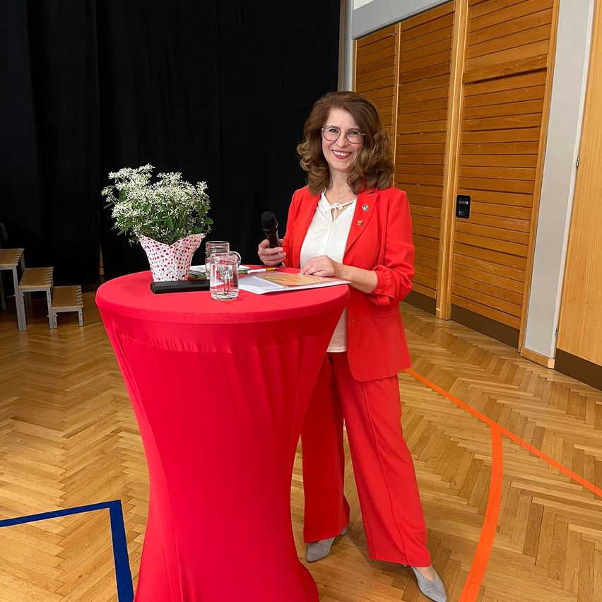 Eine Frau in einem roten Anzug steht mit einem Mikrofon an einem Tisch und lächelt. Ein Blumenvase und ein Glas Wasser sind auf dem Tisch. Hinter ihr befindet sich ein schwarzer Vorhang und eine Holzwand.