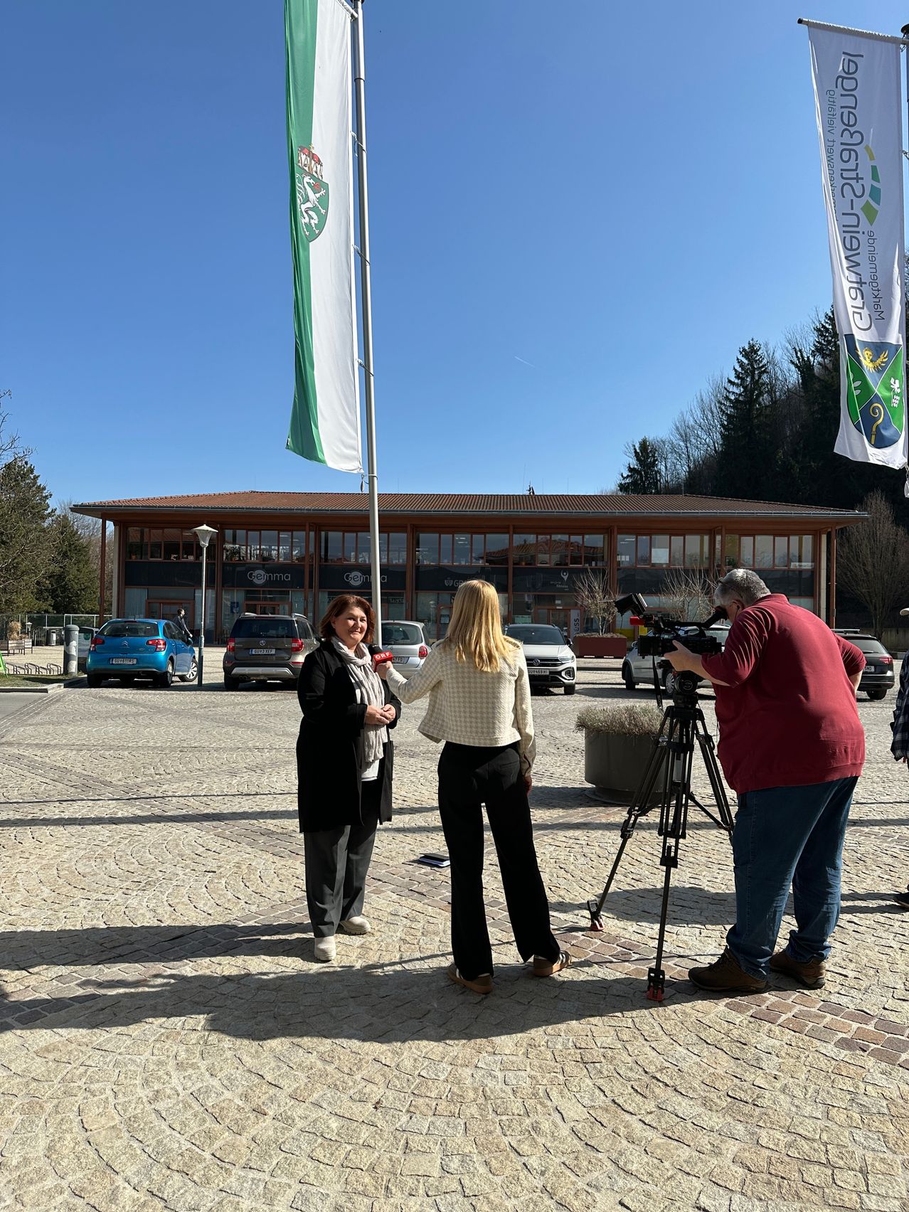 Zwei Frauen und ein Mann stehen auf einem gepflasterten Weg und werden möglicherweise interviewt. Hinter ihnen befindet sich ein Gebäude mit Fahnen. Mehrere Autos sind davor geparkt. Der Himmel ist klar.