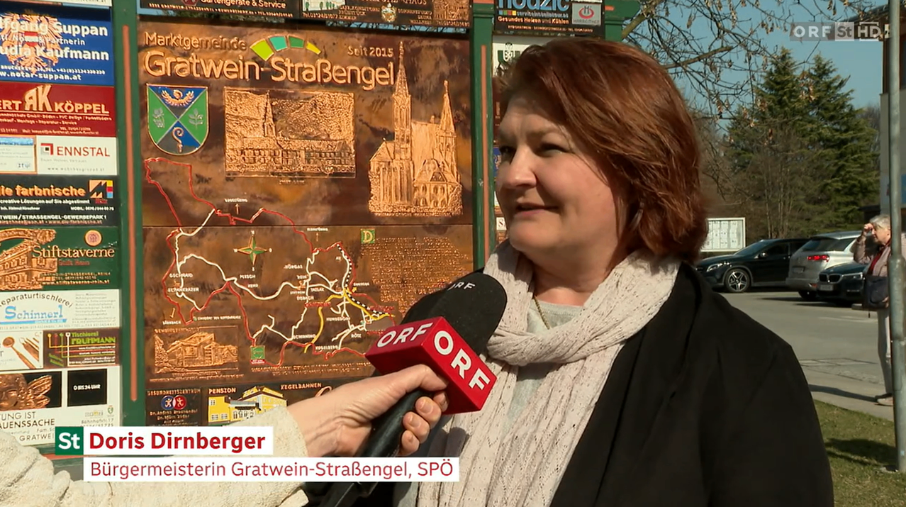Eine Frau in Gratwein-Strassengel hält ein Mikrofon mit der Aufschrift ORF und spricht in die Kamera. Hinter ihr zeigt eine Karte die Region Wein-Strassengel, mit einem Schild, das angibt, dass sie seit 2015 aktiv ist.