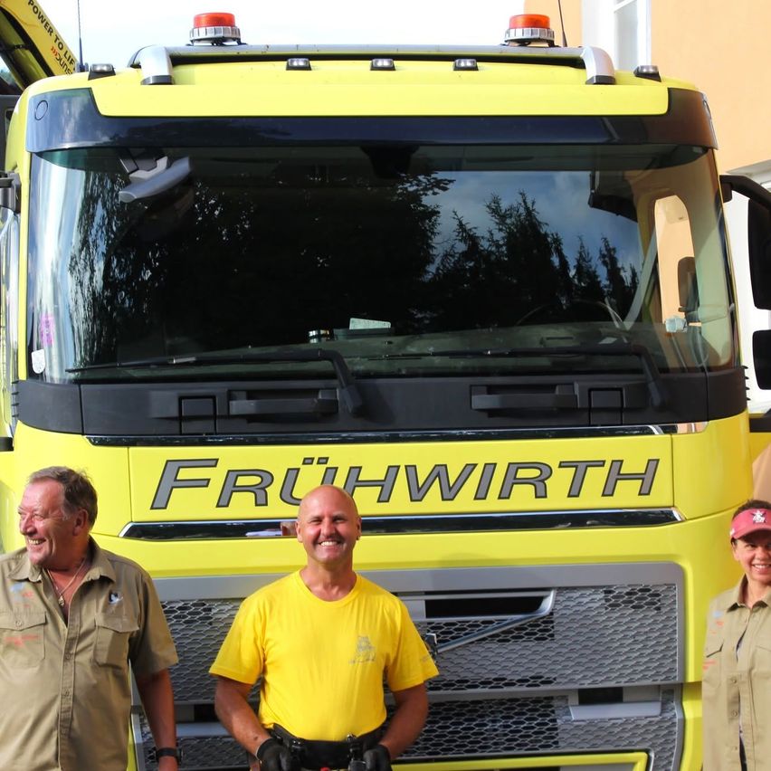Drei lächelnde Männer stehen vor einem gelben LKW mit der Aufschrift 'FRUHWIRTH'. Der Mann in der Mitte trägt ein gelbes Shirt, die anderen tragen helle Hemden.