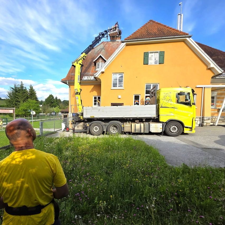 Ein Mann beobachtet, wie ein gelber Kranwagen ein Objekt vom Dach eines gelben Hauses hebt.