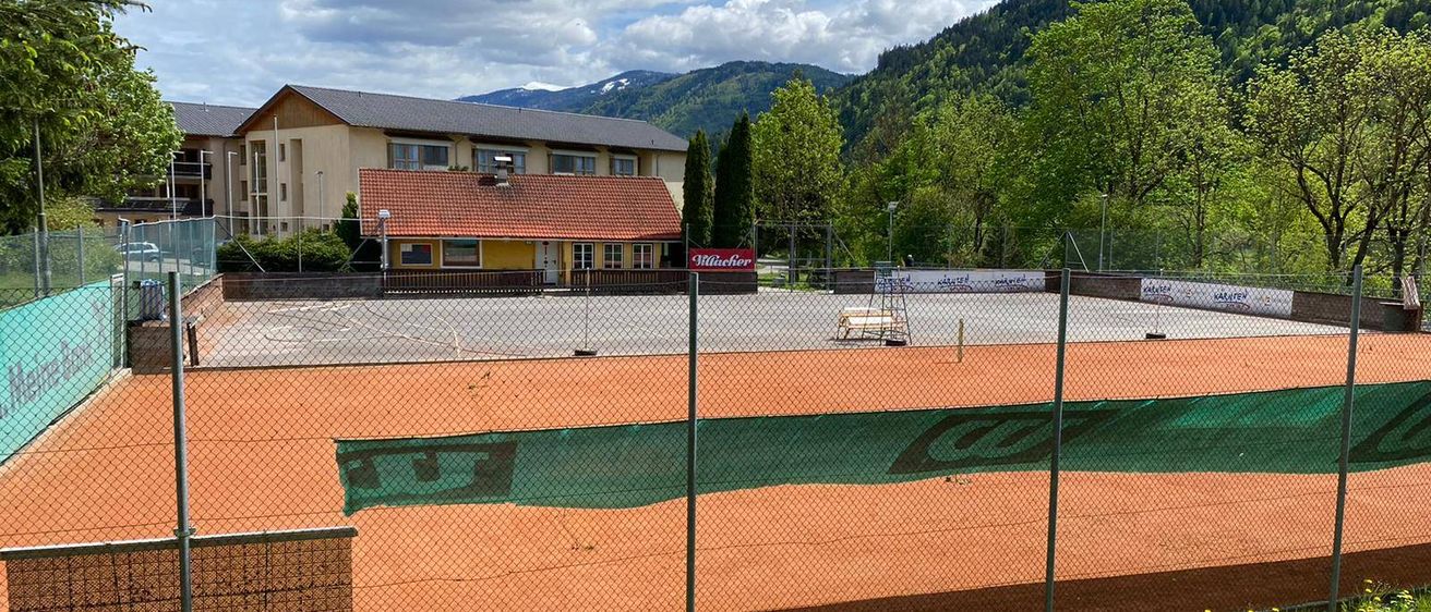 Ein leerer, eingezäunter Tennisplatz vor einem Gebäude mit rotem Dach unter einem blauen Himmel mit verstreuten Wolken. Berge sind im Hintergrund.