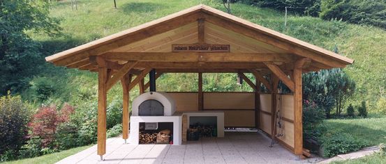 Ein Holzpavillon mit Holzofen und Holzsitzbank. Das Schild steht 'reinen Natursauer gebacken'. Grüner Rasen und Pflanzen umgeben den Bereich.