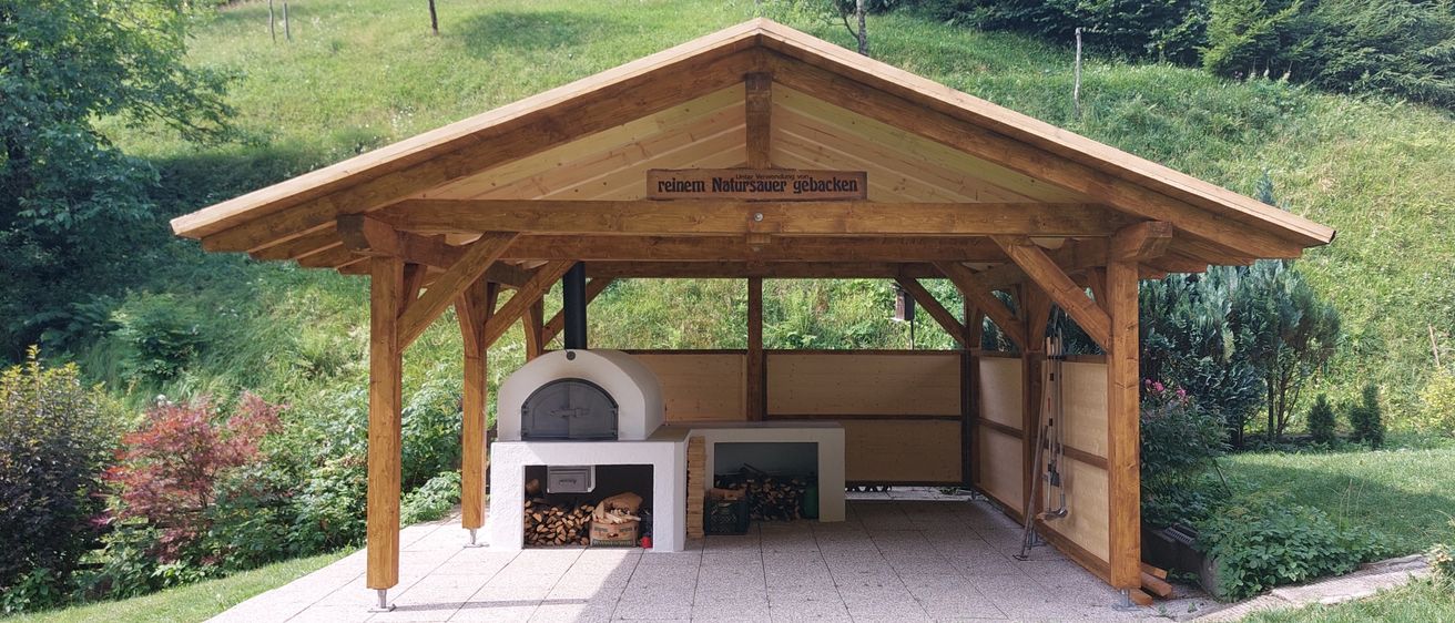 Ein Holzpavillon mit Holzofen und Holzsitzbank. Das Schild steht 'reinen Natursauer gebacken'. Grüner Rasen und Pflanzen umgeben den Bereich.