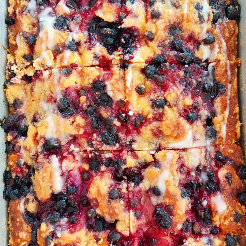 A close-up view of a blueberry cobbler with a crumbly topping, drizzled with white icing, and filled with juicy blueberries and red berry sauce.