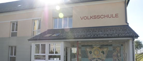 Der Eingang des Volksschule-Gebäudes zeigt eine geflieste Wand mit einem dekorativen Design. Das Gebäude hat Fenster und eine Glastür. Das Dach hat ein Dachfenster. Der Text lautet Volksschule.