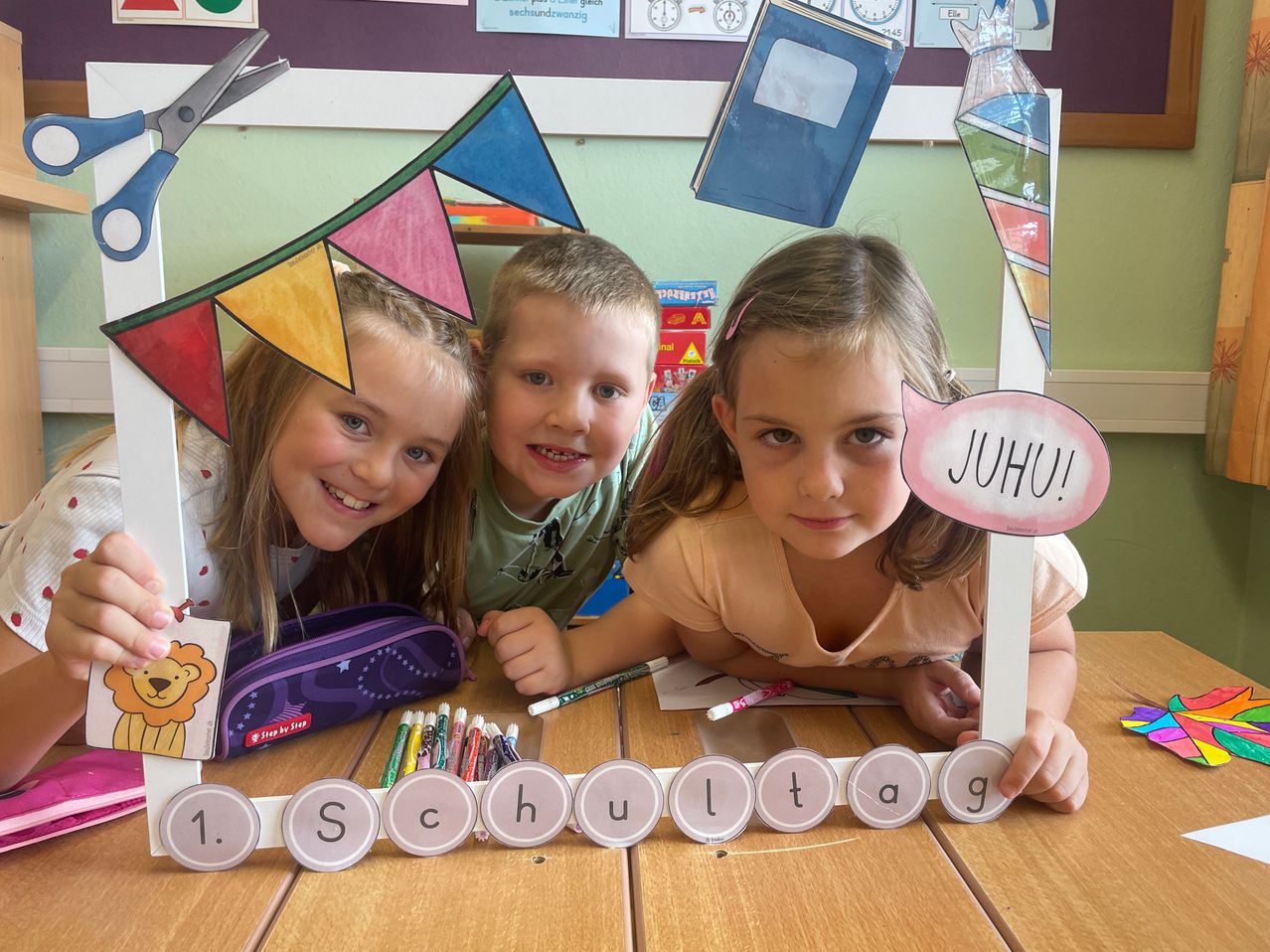 Drei Kinder im Klassenzimmer, die lächelnd für ein Foto mit einer bunten Fahne und einem Schild, das 'JUHU!' sagt, posieren. Sie sind von Schulsachen auf einem Tisch umgeben.