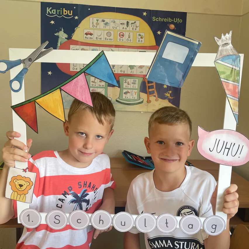 Zwei junge Jungen halten einen Fotorahmen mit bunten Dekorationen und einem Banner, auf dem '1. Schultag' steht. Sie lächeln und posieren für ein Bild. Dahinter befindet sich eine Wand mit pädagogischen Postern.