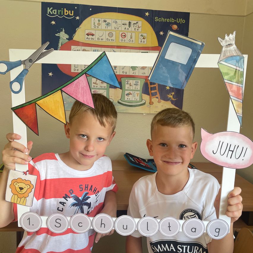 Zwei junge Jungen halten einen Fotorahmen mit bunten Dekorationen und einem Banner, auf dem '1. Schultag' steht. Sie lächeln und posieren für ein Bild. Dahinter befindet sich eine Wand mit pädagogischen Postern.