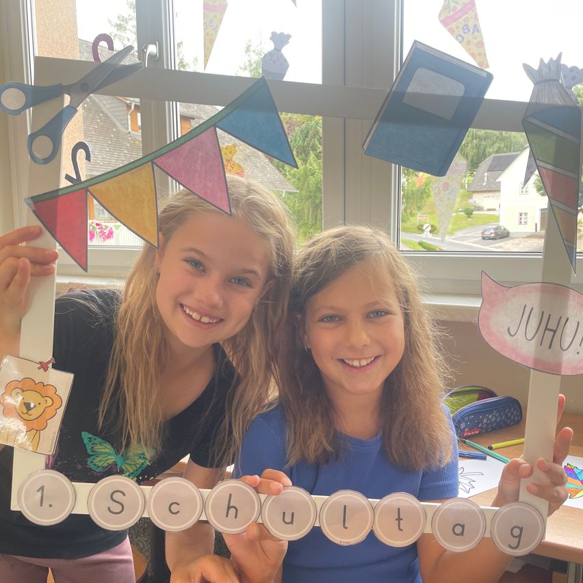 Zwei junge Mädchen lächeln und posieren für ein Foto mit einem bunten Banner, das 'Schultag' sagt. Sie halten einen Rahmen mit den Buchstaben des Wortes 'Schultag'. Hinter ihnen ist ein Fenster mit Dekorationen und Blick auf ein Haus und ein Auto.