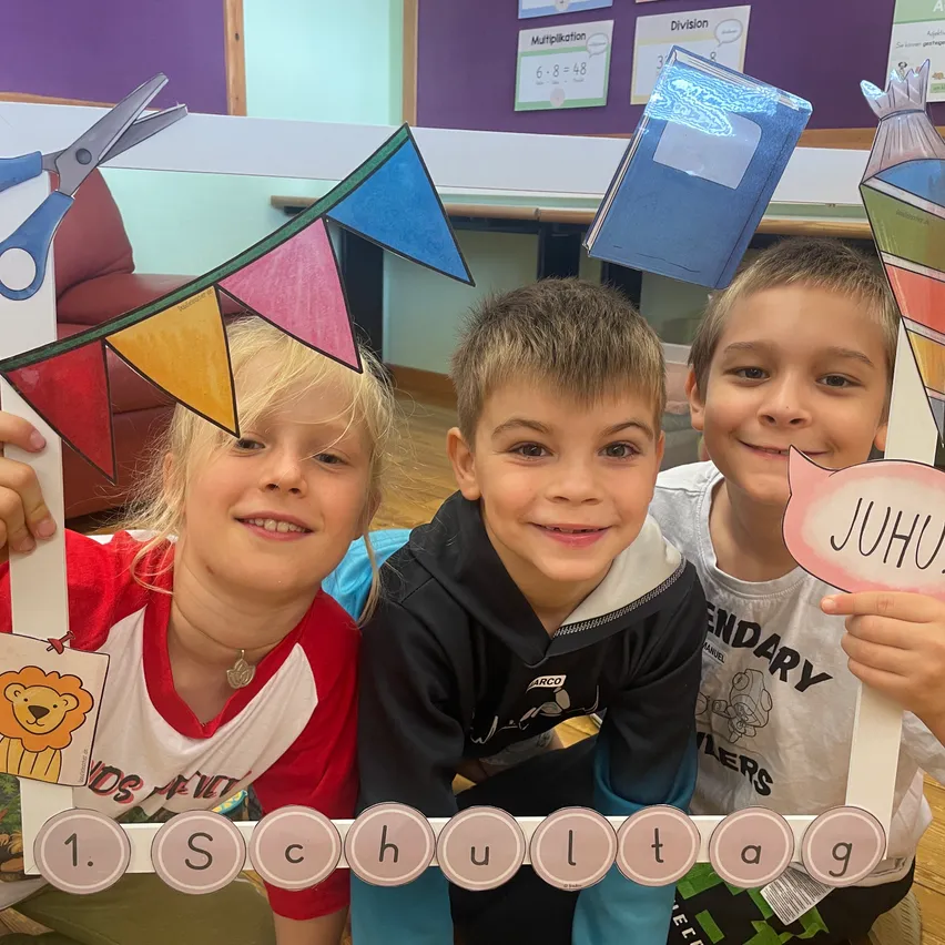 Drei Kinder lächeln und posieren für ein Foto in einer Klassenzimmerumgebung. Sie halten einen Banner mit den Worten '1. Schultag' und einem bunten Löwenausschnitt. Hinter ihnen befinden sich verschiedene pädagogische Poster und ein Schreibtisch mit Büchern.