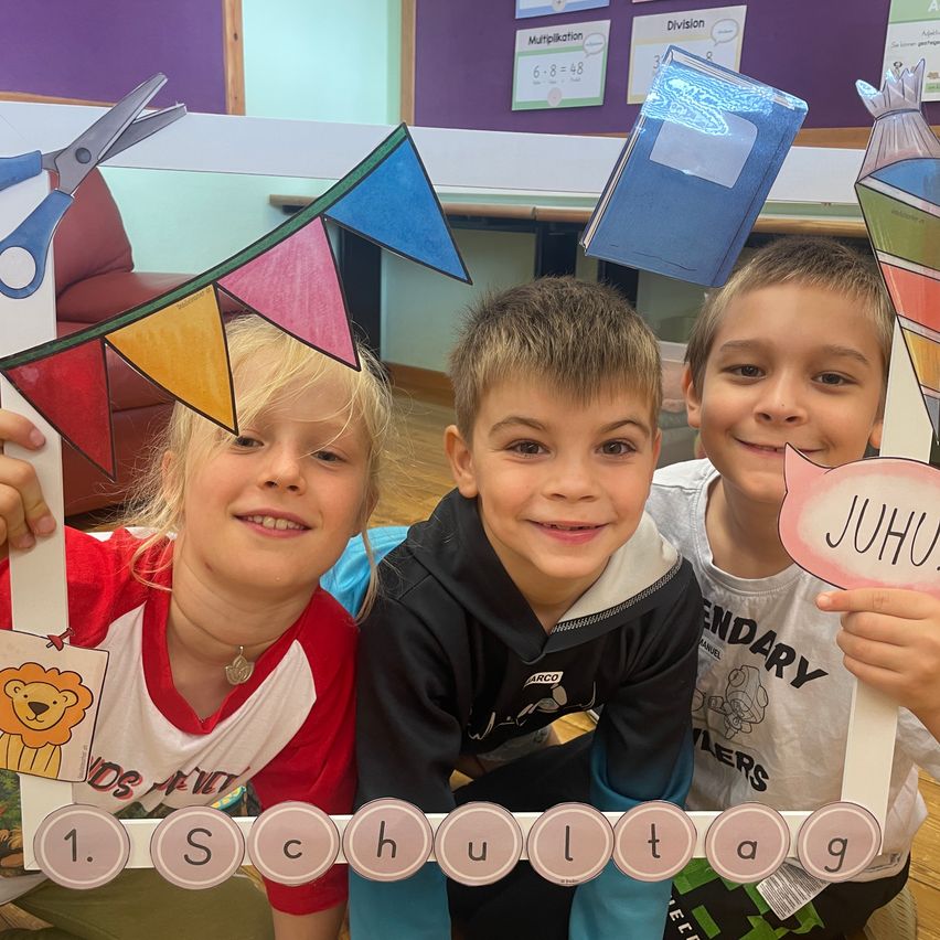 Drei Kinder lächeln und posieren für ein Foto in einer Klassenzimmerumgebung. Sie halten einen Banner mit den Worten '1. Schultag' und einem bunten Löwenausschnitt. Hinter ihnen befinden sich verschiedene pädagogische Poster und ein Schreibtisch mit Büchern.