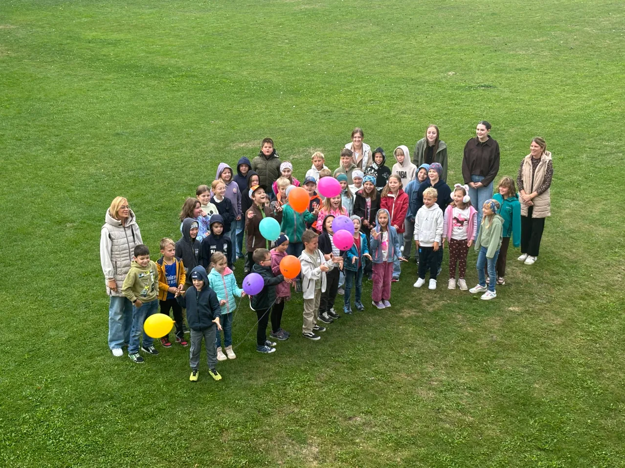 Eine große Gruppe von Kindern und Erwachsenen versammelt sich auf einem Rasenfeld und hält bunte Luftballons. Einige tragen Jacken und Turnschuhe. Die Erwachsenen stehen hinten, die Kinder vorne.