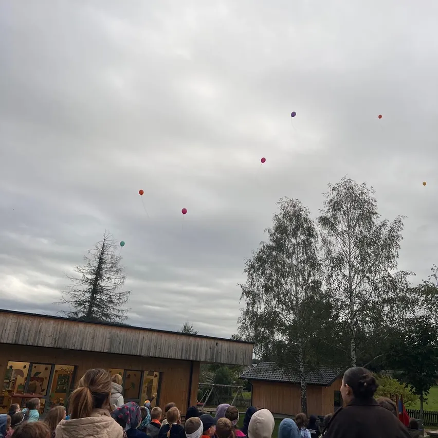 Eine Gruppe von Menschen versammelt sich draußen und beobachtet bunte Luftballons, die in der Luft schweben. Der Himmel ist bewölkt, und es gibt Bäume und ein Gebäude im Hintergrund.