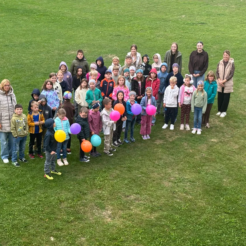 Eine Gruppe von Kindern und Erwachsenen versammelt sich auf einem Rasenfeld und posiert für ein Foto. Einige von ihnen halten bunte Luftballons.