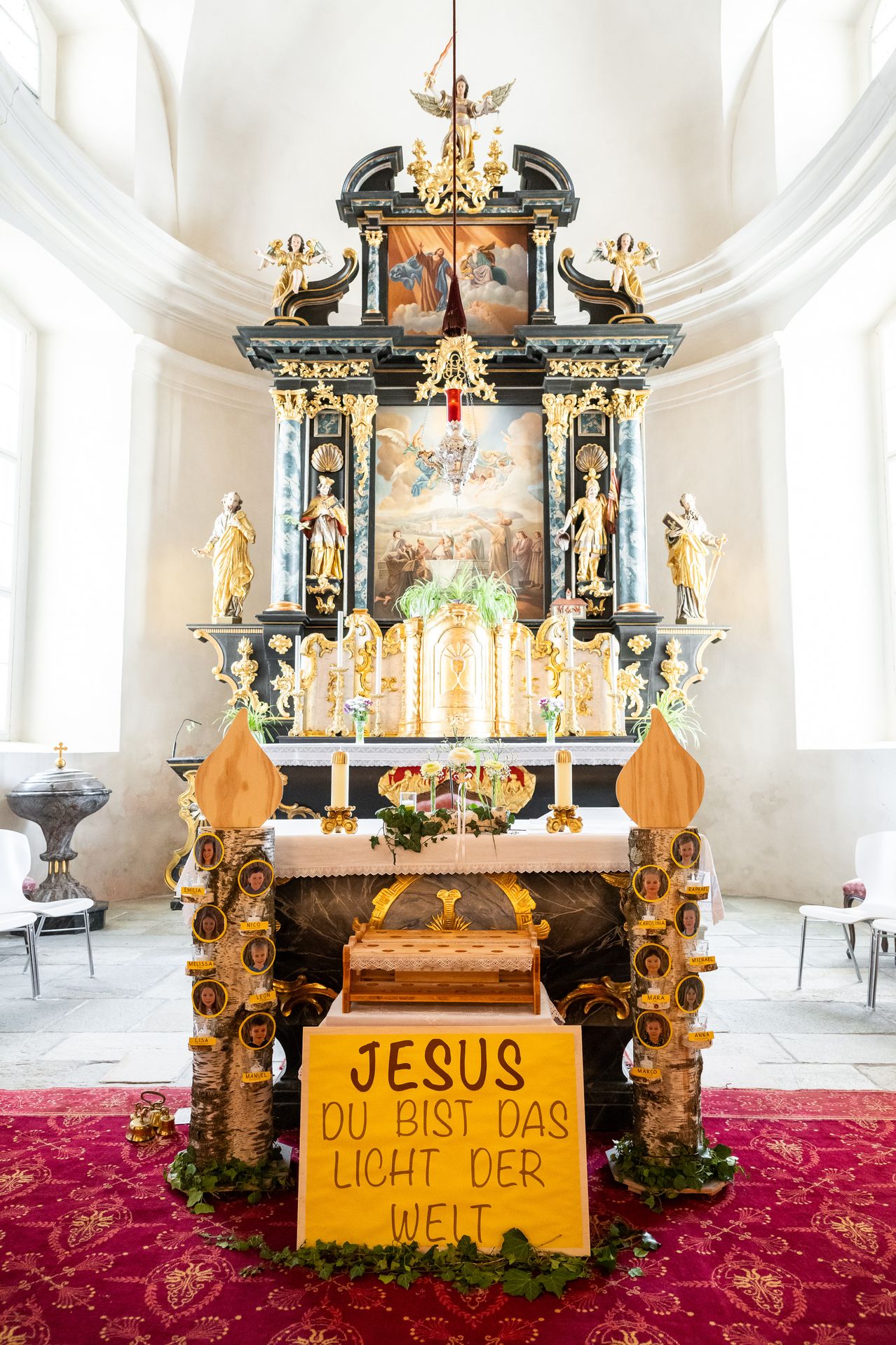 Ein Kirchenaltar mit goldenem Design und einem Gemälde dahinter. Eine Kerze ist auf dem Altar angezündet. Es gibt zwei weiße Stühle auf beiden Seiten. Ein Foto von Jesus ist in der Mitte des Altars platziert.