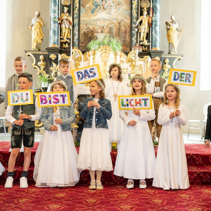 Kinder in weißen Kleidern halten Schilder mit den Worten 'Du bist Licht' und 'Das' in einer Kirche.