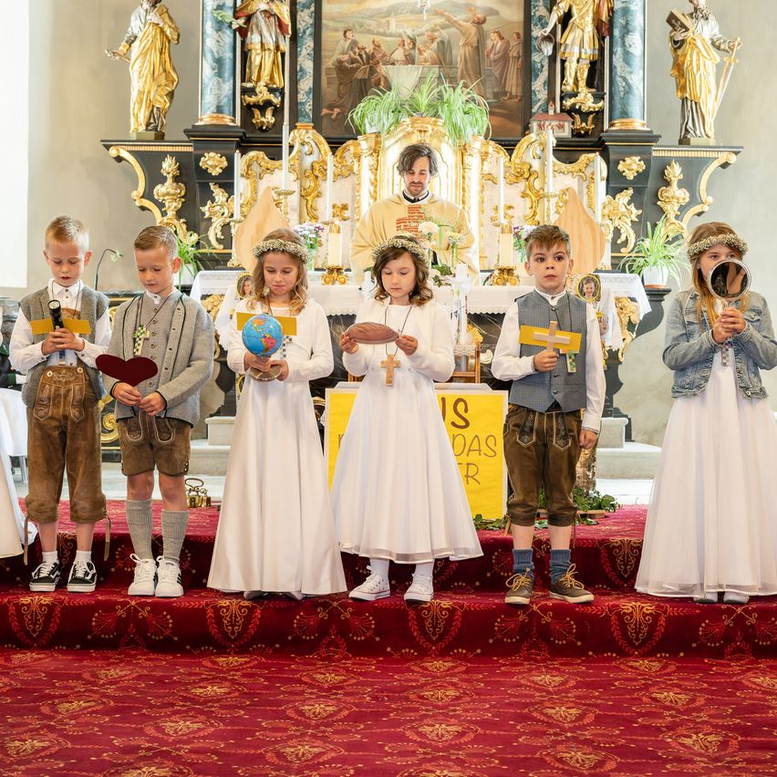 Kinder in traditionellen Kleidungsstücken stehen auf einer Bühne vor einem Kirchenaltar und halten Gegenstände wie eine Weltkugel und ein Kreuz.