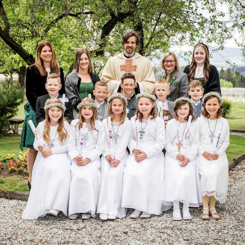Eine Gruppe von Kindern und Erwachsenen in Weiß, die in einem Garten stehen. Ein Priester in einer beigefarbenen Robe steht hinter ihnen. Einige halten Blumen.