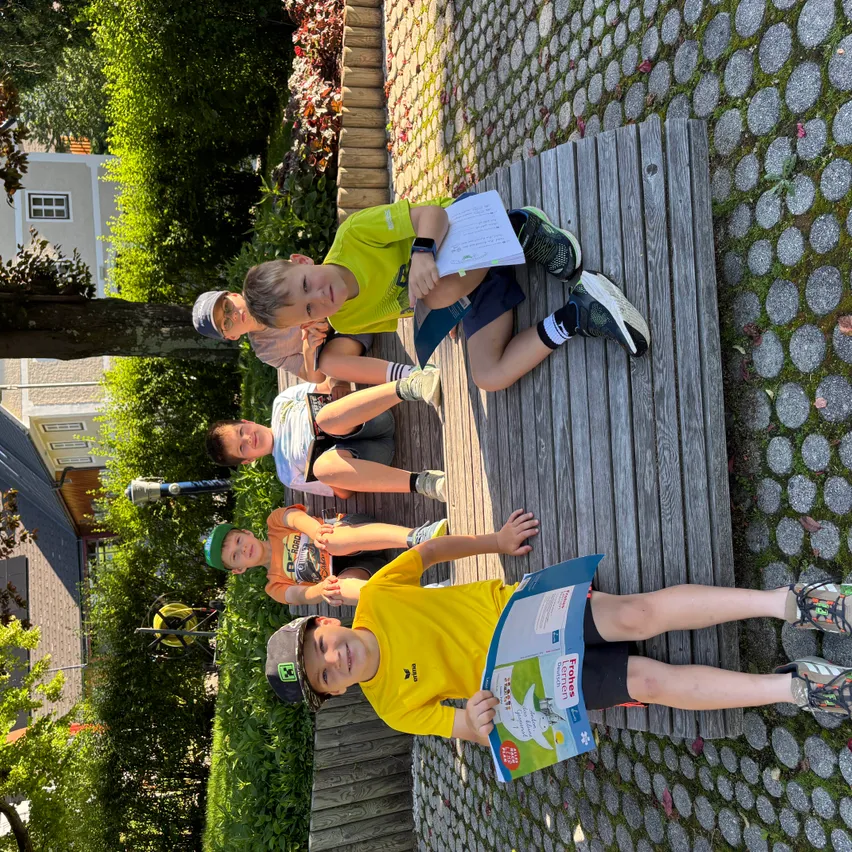 Vier Jungen in gelben T-Shirts sitzen auf einer Holzbank in einem Park und lesen Bücher. Dahinter sind Pflanzen, ein Baum und ein Gebäude.