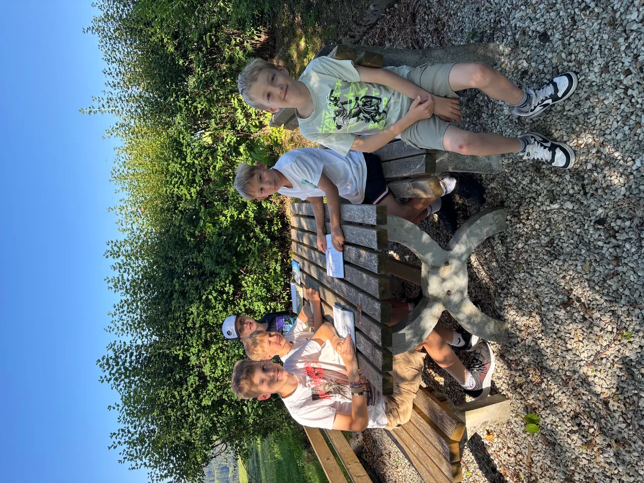 Vier Jungen sitzen auf einer Holzbank und lächeln in die Kamera. Ein Junge liest ein Papier, während andere zuschauen, mit einem Baum dahinter.