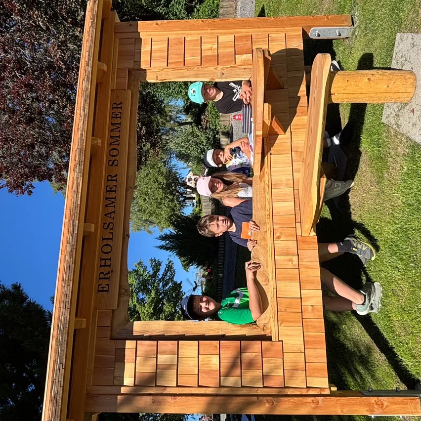 Fünf Personen sitzen in einer hölzernen Struktur mit der Aufschrift Erholsamer Sommer. Sie lächeln und posieren für ein Foto.