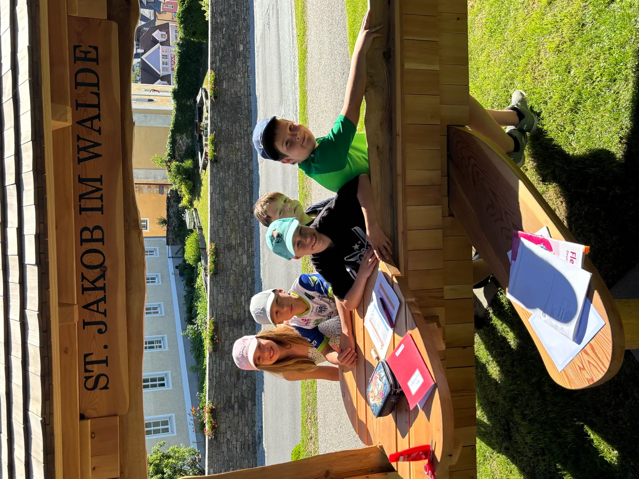 Vier Kinder lehnen sich an eine Holzstruktur in St. Jakob im Walde. Sie tragen Mützen und lächeln. Im Hintergrund befindet sich eine Straße und ein Gebäude.