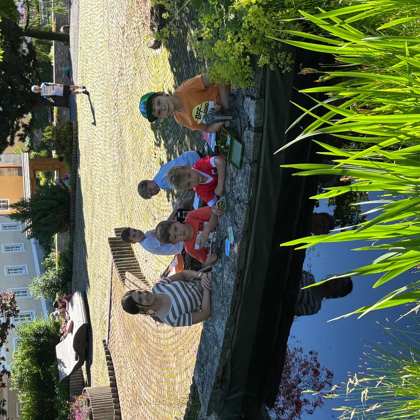 Eine Gruppe von Kindern und einer Frau sitzen in der Nähe eines Kanals und schauen sich etwas an. Der Kanal ist von Pflanzen und Bäumen umgeben. Im Hintergrund befindet sich ein Gebäude.