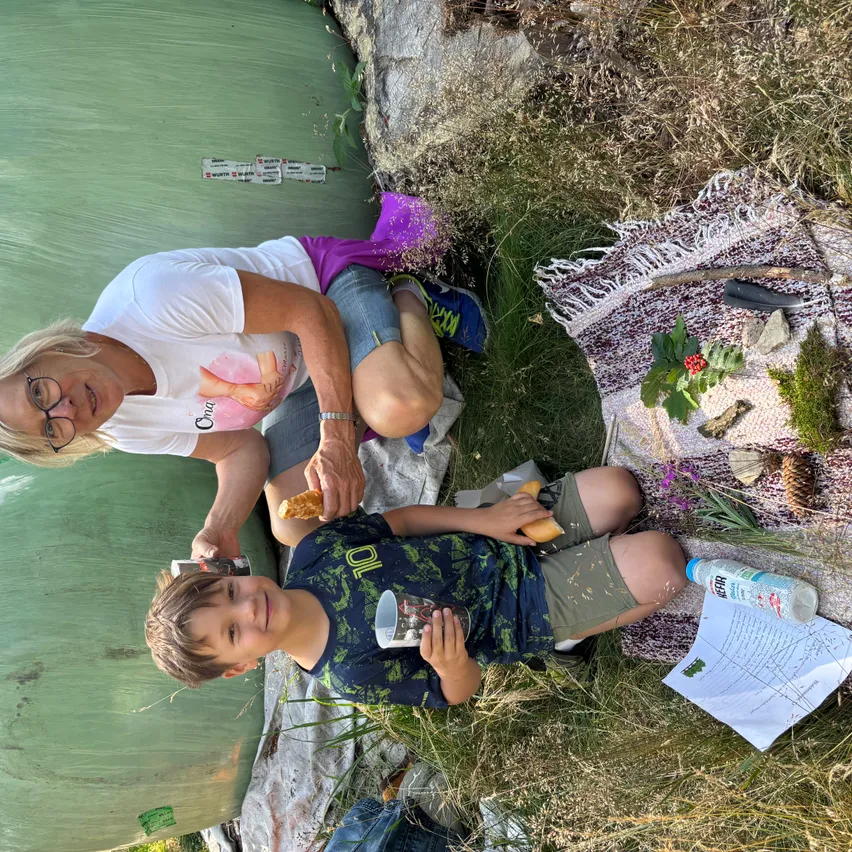 Eine Frau und ein Junge sitzen auf dem Gras und essen Sandwiches. Die Frau trägt ein weißes T-Shirt mit rosa Aufdruck und eine Brille. Der Junge hat ein blaues Shirt mit grünem Design und hält einen Becher.