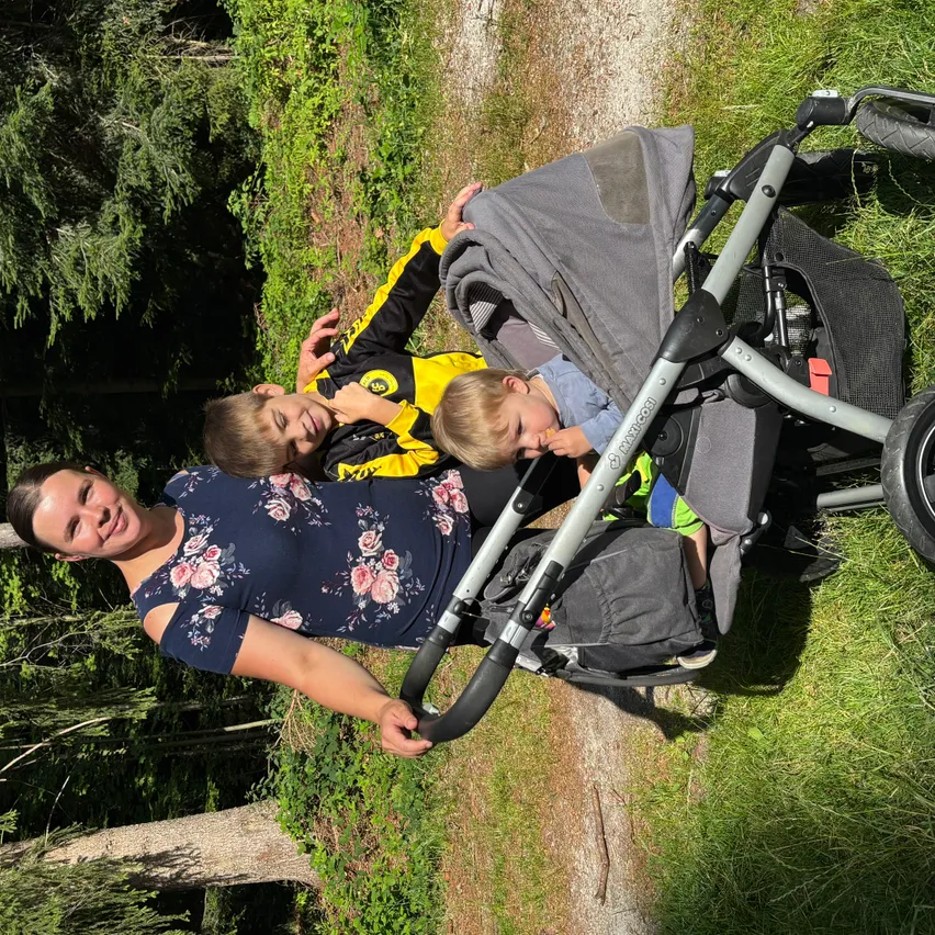 Eine Frau und zwei junge Jungen sind draußen, die Frau schiebt einen Kinderwagen, in dem ein Junge sitzt, während der andere Junge neben ihr steht und lächelt.
