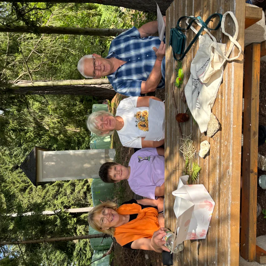 Vier Personen sitzen an einem Picknicktisch im Wald und lächeln. Ein Mann und eine Frau stehen hinter dem Tisch, während zwei Kinder davor sitzen. Eine Tasche, eine Handtasche und andere Gegenstände sind auf dem Tisch. Bäume sind hinter ihnen und eine Struktur ist auf der linken Seite zu sehen.