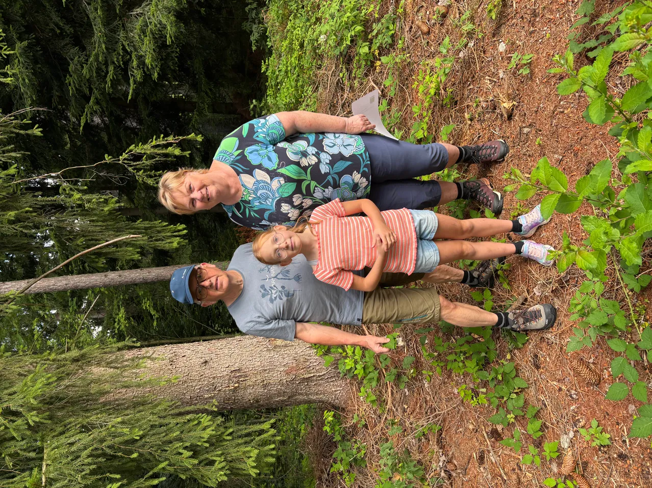 Eine Familie aus drei Personen, bestehend aus einer Frau, einem Mann und einem Kind, liegt auf dem Waldboden. Die Frau hält ein Stück Papier.