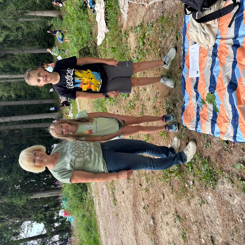 Eine Gruppe von Menschen versammelt sich in einem Waldgebiet, mit einer Decke und einem Rucksack auf dem Boden. Ein Junge und ein Mädchen stehen, während eine ältere Frau sitzt. Bäume umgeben das Gebiet.