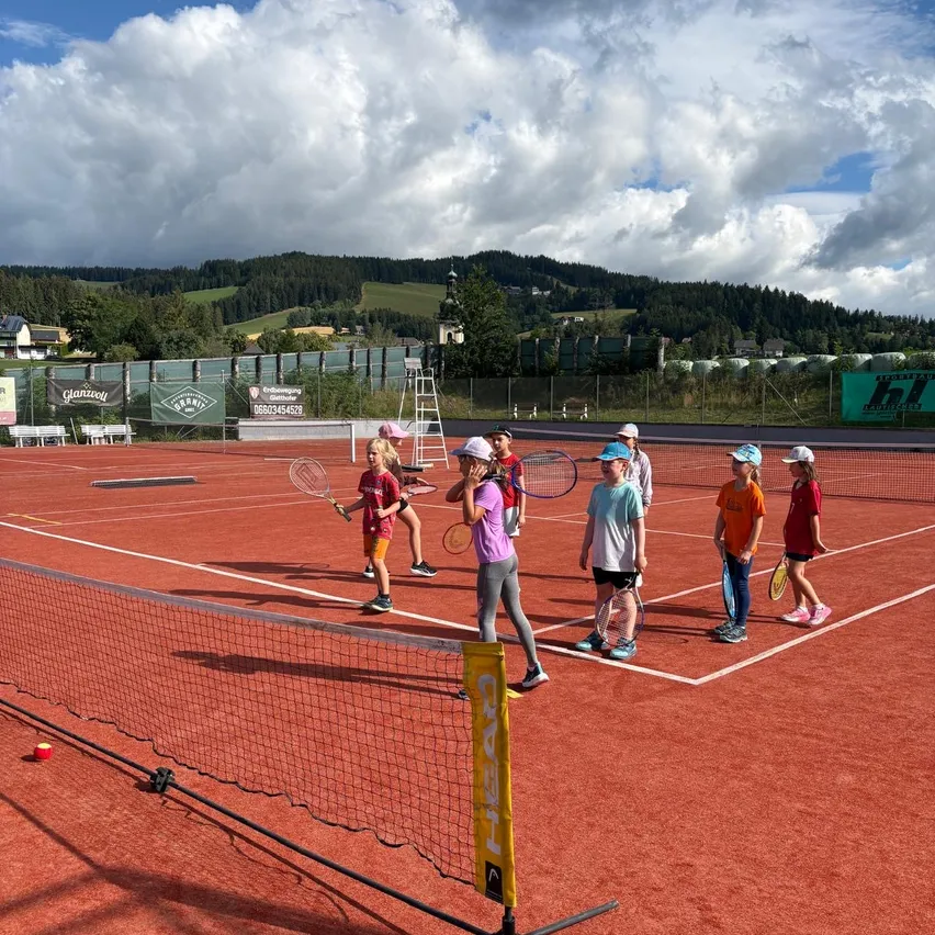 Eine Gruppe von Kindern trainiert Tennis auf einem roten Platz, mit Hügeln und Bergen im Hintergrund.