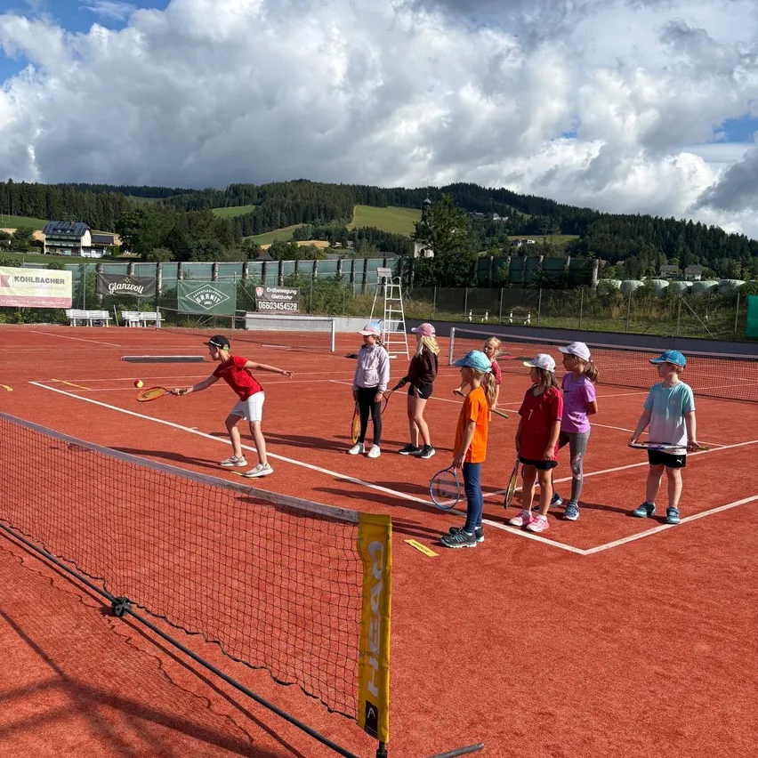 Kinder üben Tennis auf einem rotbeschichteten Platz, umgeben von einem Zaun. Ein Kind schlägt einen Ball, während andere zusehen. Im Hintergrund befinden sich Bäume und Berge.
