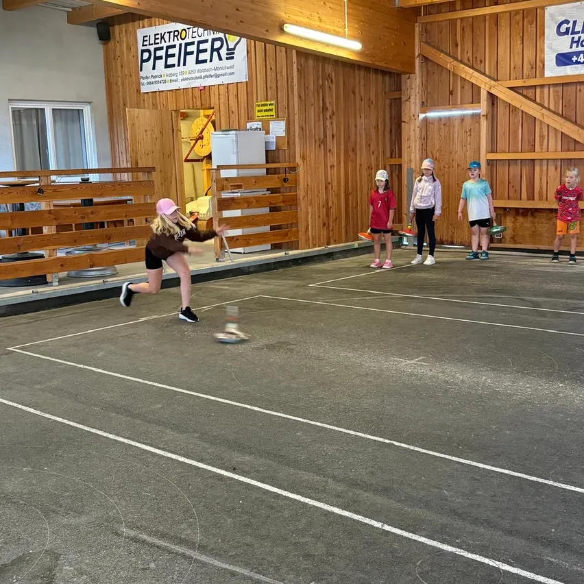Ein Mädchen wirft einen Ball auf einem Spielfeld, während vier Kinder zuschauen. Das Spielfeld hat weiße Linien. Hinter den Kindern befinden sich Holzzäune und ein Schild.