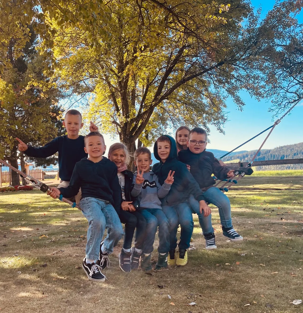 Eine Gruppe von Kindern, einige sitzend und einige stehend, posiert für ein Foto unter einem Baum mit gelben Blättern. Sie alle lächeln und machen Friedenszeichen.
