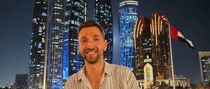 Ein Mann lächelt in die Kamera und steht vor einer nächtlichen Stadtlandschaft. Hinter ihm sind mehrere Hochhäuser mit blauen und weißen Lichtern beleuchtet. In der Ferne ist eine Fahne auf einem Fahnenmast zu sehen.