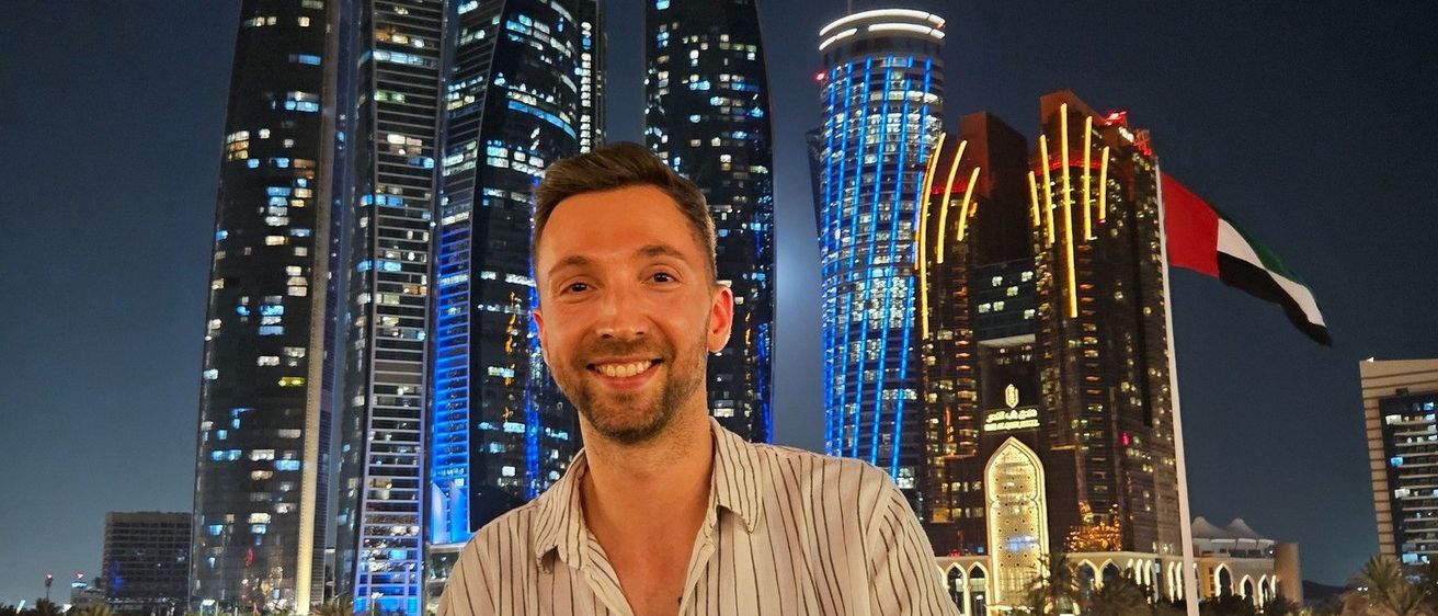 Ein Mann lächelt in die Kamera und steht vor einer nächtlichen Stadtlandschaft. Hinter ihm sind mehrere Hochhäuser mit blauen und weißen Lichtern beleuchtet. In der Ferne ist eine Fahne auf einem Fahnenmast zu sehen.