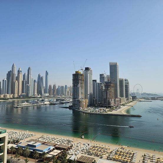 Ein Luftbild einer Stadt mit vielen Hochhäusern, einem Strand und einem Riesenrad. Die Stadt hat einen großen Wasserkörper mit Booten.