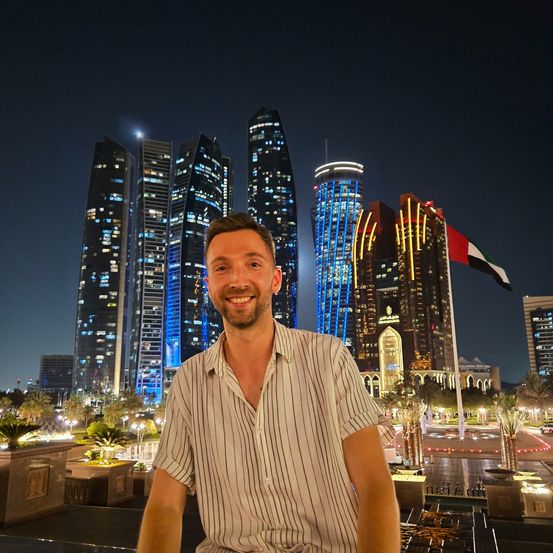 Ein lächelnder Mann steht bei Nacht vor der Skyline von Abu Dhabi, mit wehender UAE-Flagge im Hintergrund. Hohe Gebäude mit leuchtenden Lichtern bilden einen lebendigen urbanen Hintergrund.
