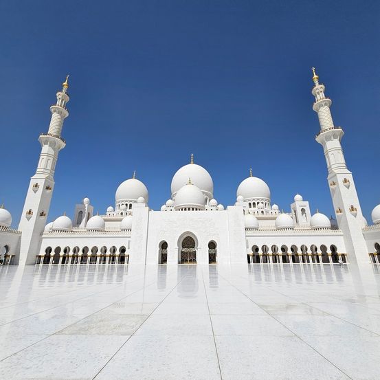 Die Sheikh-Zayed-Großmoschee in Abu Dhabi verfügt über zwei hohe Minarette, zahlreiche Kuppeln und einen großen Eingang, die sich alle im Marmorhof unter einem klaren blauen Himmel widerspiegeln.
