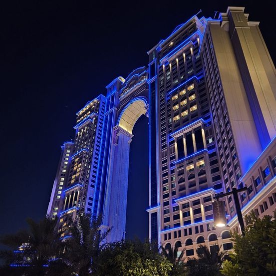 Ein großes Gebäude mit blauen Lichtern in der Nacht. Es hat einen hohen Bogen und eine zentrale Säule. Viele Fenster sind beleuchtet.