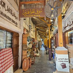 Eine Marktgasse mit Holzregalen, die verschiedene geflochtene Körbe zeigen. Die Decke hat Ventilatoren und Lampen. Ein Schild steht Utensil Souq. Eine Person sitzt auf einer Bank, und Kisten sind rechts gestapelt.